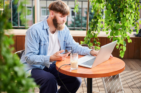 Man works on laptop and smartphone at outdoor table, with glass of iced coffee. Solar panel nearby, emphasizing sustainable, modern eco-friendly workspace that combines technology and sustainability.の写真素材