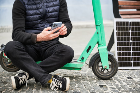 Cropped view of man sits on electric scooter with solar panel and smartphone while charging. Integration of renewable energy in personal transportation, highlighting sustainable urban lifestyle.の写真素材