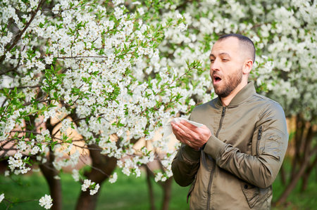 Man allergic suffering from seasonal allergy at spring in blossoming garden at springtime. Man sneezing and blowing nose using nasal handkerchief in front of blooming tree. Spring allergy concept.の写真素材