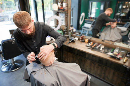 Man barber cutting little boys hair using comb and scissors. Child getting haircut from adult male, likely barber. Professional hairdresser and cute client at modern barbershop.の写真素材