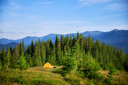 Bright yellow tourist tent on grassy hillside, surrounded by lush green pine trees. Breathtaking view of distant blue mountains under clear sky, creating idyllic and serene camping spot.の写真素材