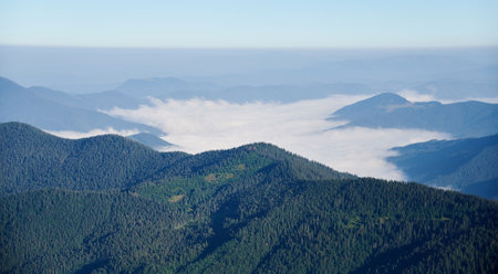 Vast mountain landscape under clear blue sky. Layers of forested hills and peaks stretch into distance, with valleys filled with sea of clouds. Lush greenery of trees contrasts with soft white clouds.の写真素材
