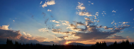 Serene evening sky with hues of blue and soft pink clouds as sun sets. Horizon glows gently, casting light over silhouetted pine trees and rolling hills, creating peaceful and expansive landscape.の写真素材