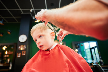 Man barber cutting little boys hair using comb and scissors. Child getting haircut from adult male, likely barber. Professional hairdresser and cute client at modern barbershop.の写真素材