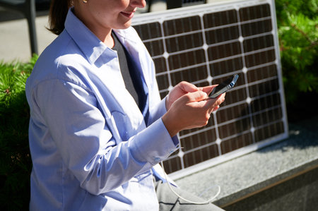 Happy woman using smartphone connected to solar panel. Cropped view. Integration of sustainable renewable energy into everyday life, demonstrating practical use of solar power for charging devices.の写真素材