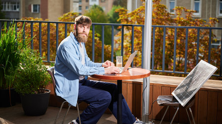Bearded man works on laptop at outdoor round table, with glass of iced coffee. Solar panel nearby, emphasizing sustainable, modern eco-friendly workspace that combines technology and sustainability.の写真素材