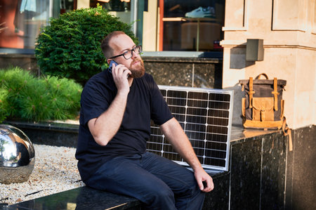 Bearded man talks on smartphone next to photovoltaic solar panel. Integration of sustainable renewable energy into everyday life, demonstrating practical use of solar power for charging devices.の写真素材
