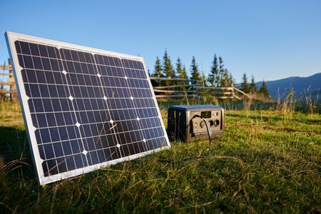 Tourist tent with solar panel and portable power station nearby in grassy field. Scene surrounded by rolling hills and wooden fence under clear blue sky, showcasing eco-friendly camping setup.の写真素材