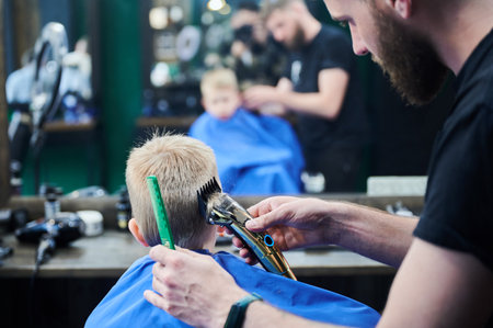 Barber using comb and shaver to cut hair. Back view of male hairdresser shaving little kids nape. Professional barber and cute boy in front of mirror.の写真素材