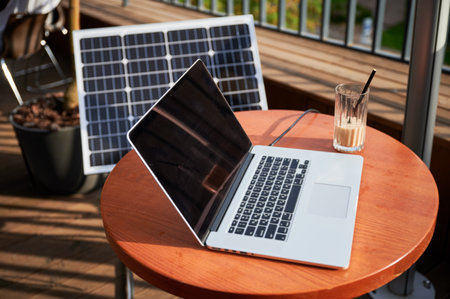 Laptop on round wooden table next to glass of iced coffee on sunny balcony. In background, solar panel propped up, symbolizing blend of technology and sustainable energy in modern workspace.の写真素材