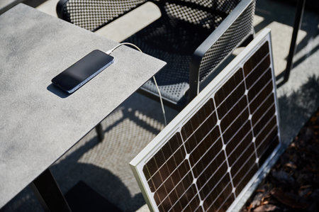 Smartphone connected to solar panel on outdoor table, emphasizing practical use of solar energy for charging devices. Integration of renewable energy, showcasing portable solar technology.の写真素材