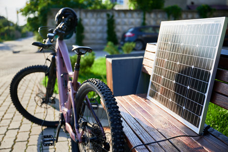 Pink electric mountain bike parked next to wooden bench, connected to solar panel for charging. Bike, with helmet hanging on handlebars, rests on paved area surrounded by greenery and grass.の写真素材