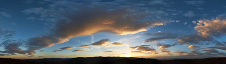Stunning sunset fills sky with rich hues of blue and orange. Dramatic clouds with hints of pink and gold spread across sky. Silhouettes of hills in foreground add depth to breathtaking evening scene.の写真素材
