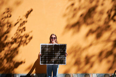 Woman holding solar panel for charging, stands against vibrant orange wall. Smiling girl wears sunglasses, white top, blue jeans. Artistic shadows cast by nearby trees branches on wall.の写真素材