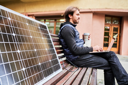 Man sits on wooden bench in urban setting, working on laptop with cup of coffee in hand. Solar panel rests beside, emphasizing sustainable, mobile workspace amidst backdrop of historic architecture.の写真素材