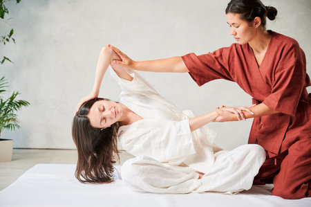 Masseuse making thai yoga massage. Woman therapist doing traditional massage treatment, stretching female patients body. Client sitting on mat, enjoying hand massage in calm atmosphere.の写真素材