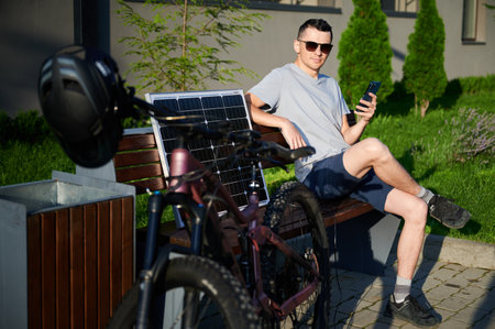 Man cyclist using solar panel for charging electric mountain bike outdoors. Guy in sunglasses sits on bench, holding phone, casually dressed in t-shirt, shorts, enjoying sunny outdoor setting.の写真素材