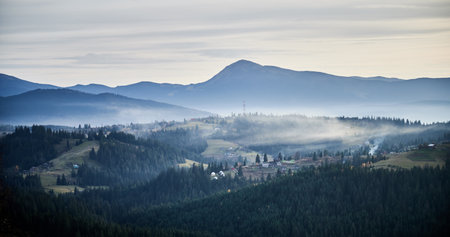 Misty mountain landscape at dawn, with layers of dark green forests and rolling hills. Small rural homes and communication tower scattered across hillsides. Distant mountains bathed in soft light.の写真素材
