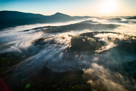 Stunning aerial view of sunrise over mountainous landscape, with thick fog blanketing valleys and hills. Suns golden rays pierce through mist, casting serene glow over the entire scene.の写真素材