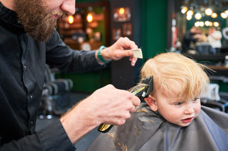 Professional hairdresser shaving boys head, using shaver and comb. Cute boy excited about new haircut. Blond boy getting stylish haircut in barbershop.の写真素材