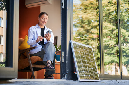 Happy woman using smartphone connected to photovoltaic solar panel. Integration of sustainable renewable energy into everyday life, demonstrating practical use of solar power for charging devices.の写真素材