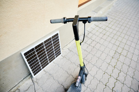 Electric scooter with yellow handlebar parked on patterned sidewalk next to beige wall. Solar panel leans against wall, connected to scooter, highlighting clean energy charging setup in urban setting.の写真素材