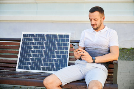 Handsome man using smartphone connected to photovoltaic solar panel. Integration of sustainable renewable energy into everyday life, demonstrating practical use of solar power for charging devices.の写真素材