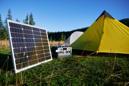 Tourist tent with solar panel and portable power station nearby in grassy field. Scene surrounded by...