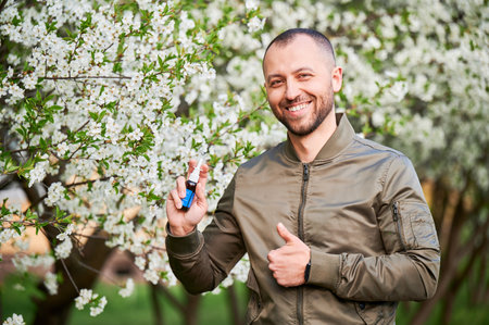 Man allergic using medical nasal spray, suffering from seasonal allergy at spring in blossoming garden. Portrait of smiling man showing thumbs up near blooming tree outdoors. Spring allergy concept.の写真素材