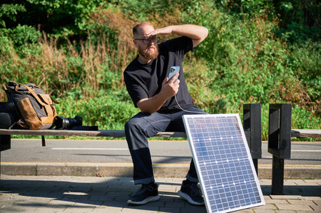 Bearded man using smartphone connected to photovoltaic solar panel. Integration of sustainable renewable energy into everyday life, demonstrating practical use of solar power for charging devices.の写真素材