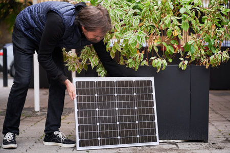 Man crouches down to place solar panel near large planter with green foliage. Guy in black outfit and navy blue quilted vest. Concept on integrating sustainable energy with city life.の写真素材