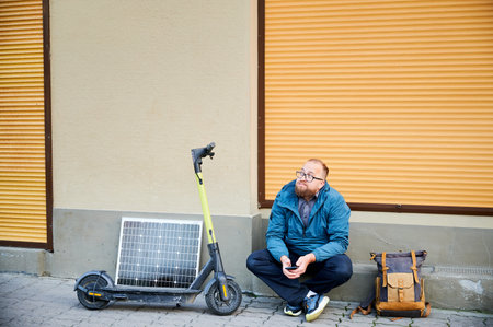 Male person waits while charging electric scooter with photovoltaic solar panel in urban settings. Integration of solar power as sustainable energy source, promoting eco-friendly urban transportation.の写真素材