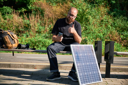 Bearded man using smartphone connected to photovoltaic solar panel, giving thumbs up. Integration of sustainable renewable energy into everyday life, practical use of solar power for charging devices.の写真素材