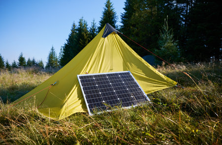 Yellow tent with solar panel set up in grassy field surrounded by dense evergreen trees under clear blue sky. Eco-friendly camping, integration of renewable energy in outdoor activities.の写真素材