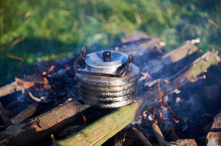 Close-up of metal kettle heating over campfire. Flames and smoke curl around well-used kettle, which rests on logs. Blur of green grass, emphasizing rustic outdoor setting on background.の写真素材