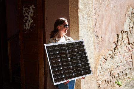 Woman holding solar panel for charging, stands against weathered, textured wall. Smiling female in sunglasses wears white top and blue jeans, standing in sunlit alley in urban scene.の写真素材