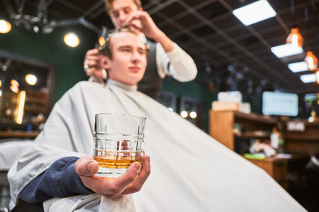 Close up photo of young client holding glass. Male customer relaxing while getting new haircut. Professional hairdresser in fancy outfit cutting clients hair.の写真素材