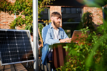 Man works on laptop at outdoor round table, with glass of iced coffee. Solar panel nearby, emphasizing sustainable, modern eco-friendly workspace that combines technology and sustainability.の写真素材
