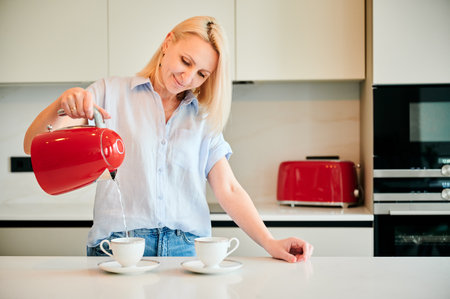 Young woman making morning coffee. Pretty female filling cup with hot water. Woman holding red kettle and pouring water into cup.の写真素材