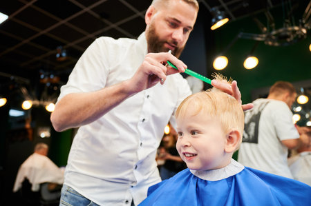 Man barber cutting little boys hair using comb and scissors. Child getting haircut from adult male, likely barber. Professional hairdresser and cute client at modern barbershop.の写真素材
