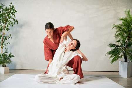 Masseuse making thai yoga massage. Woman therapist doing traditional massage treatment, stretching female patients body. Client sitting on mat, enjoying hand massage in calm atmosphere.の写真素材