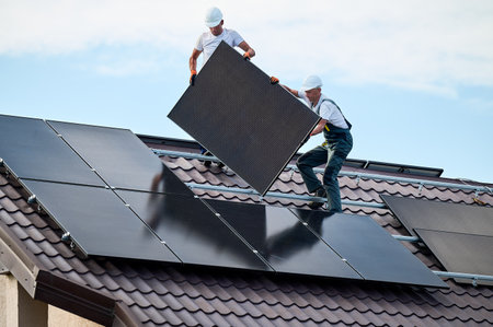 Workers building solar panel system on roof of house. Two men installers in helmets carrying photovoltaic solar module outdoors. Alternative, green and renewable energy generation concept.の写真素材
