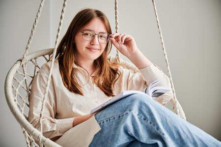 Portrait of happy girl relaxing and reading book on indoor swing. Pretty female in glasses excited about story she reading. Cozy afternoon with book in hanging chair in home.の写真素材