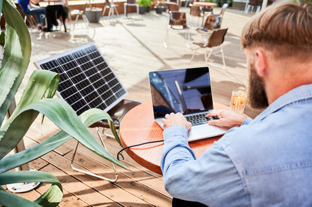 Man works on laptop at outdoor round table, with glass of iced coffee. Solar panel nearby, emphasizing sustainable, modern eco-friendly workspace that combines technology and sustainability.の写真素材