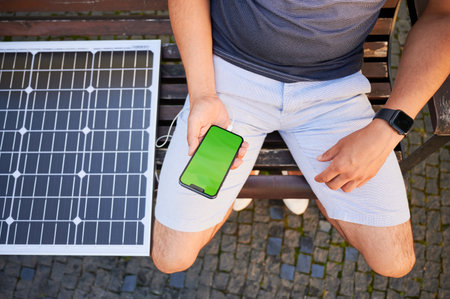 Young man using portable solar panel to charge his phone with green screen. Cropped photo of male sitting on bench and charging his mobile device by solar battery. Saving energy concept.の写真素材