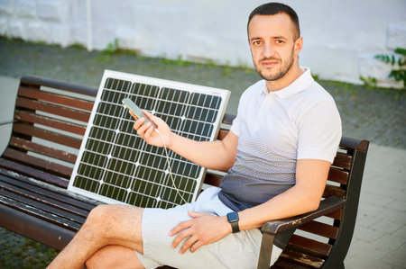 Handsome man sitting on bench while his phone charging from solar battery. Young man waiting for his smartphone to charge. Alternative energy in everyday usage.の写真素材