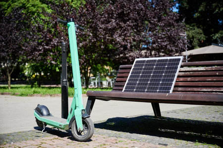 Mint-green electric scooter parked on cobblestone path next to bench in park. On bench, solar panel...