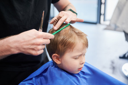 Man barber cutting little boys hair using comb and scissors. Child getting haircut from adult male, likely barber. Professional hairdresser and cute client at modern barbershop.の写真素材