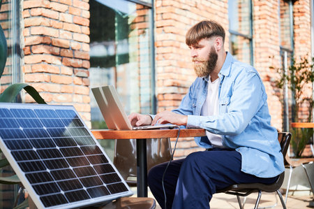 Man works on laptop at outdoor round table, with glass of iced coffee. Solar panel nearby, emphasizing sustainable, modern eco-friendly workspace that combines technology and sustainability.の写真素材