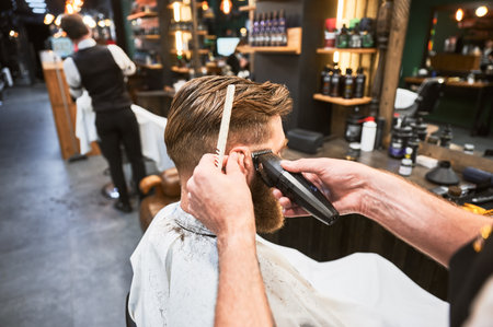 Professional barber using comb and electric shaver to cut side hair. Close-up view of male customer getting stylish haircut. Side view of man on background of other hairdresser.の写真素材